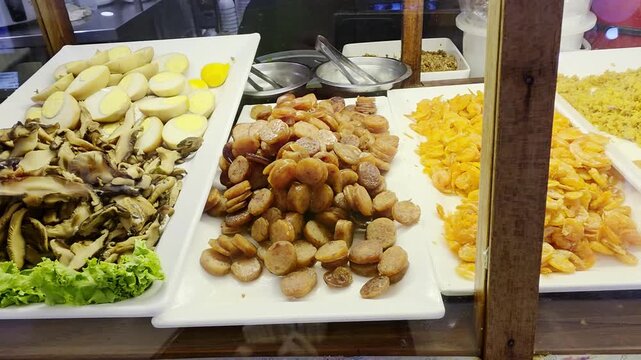 Selection of traditional Thai street food ingredients on white trays including sliced Chinese sausage, dried shrimp, and savory toppings displayed behind a glass counter at a local food stall.