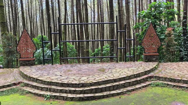 Outdoor amphitheater stage with stone floor surrounded by tall pine forest trees decorated with traditional Javanese gunungan art ornaments in a serene natural park for performance events and gatherin