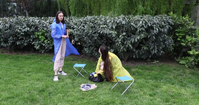 Women in raincoats gather food and items while finish picnic of bad weather. Young females interrupt outdoor resting due to rain in spring garden