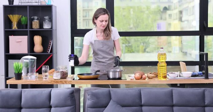 Woman in apron mixes dish in pan and adds seasoning at induction stove in kitchen. Happy housewife cooks tasty healthy dinner in studio apartment