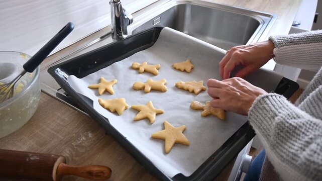 Close-up of hands arranging star and gingerbread man shaped cookie dough on a parchment lined baking tray while preparing homemade Christmas cookies in the kitchen.