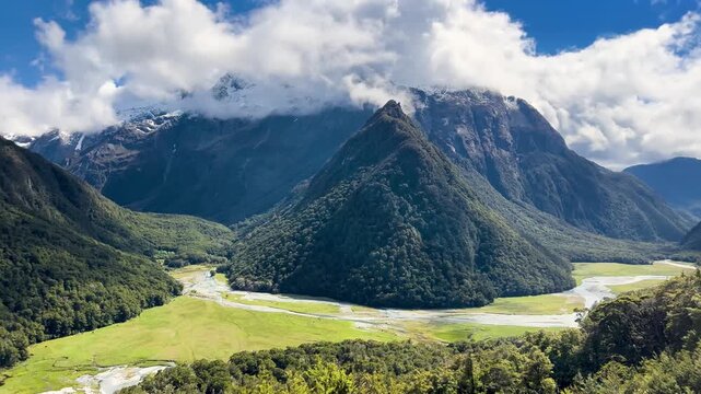 A braided river winds across the green Routeburn Flats, New Zealand, a forested pyramid of a peak standing at the valley head below snow streaked mountains. Pan right shot