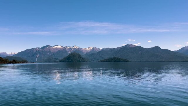 Calm water stretches across Lake Manapouri in Fiordland, New Zealand, forested islands lying low in front of a long range of snow streaked mountains. Wide shot