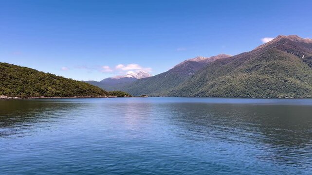 A big forested mountain drops straight into Lake Manapouri beside a low wooded headland, with pale snow on the far range under a clear sky. Wide shot