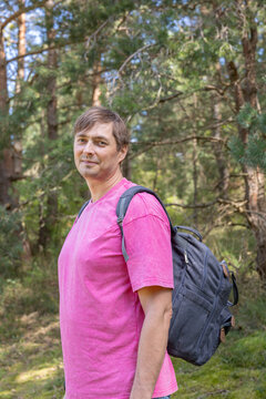 Adult man in pink T-shirt with backpack smiling and looking at camera in forest on sunny day