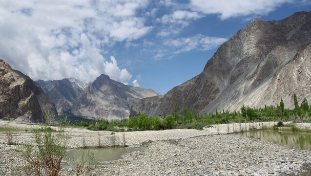 View of the rugged mountain landscape along the K2 trek with a rocky riverbed, lush green trees, and towering peaks under a blue sky in Hushe, Pakistan.