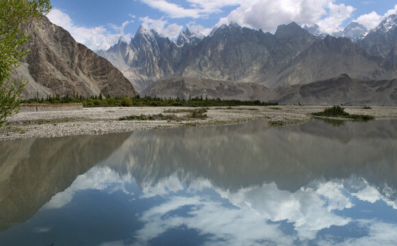 View of the K2 trek landscape in Hushe, Pakistan, featuring a calm river reflecting the dramatic snow-capped peaks and clouds under a bright sky.