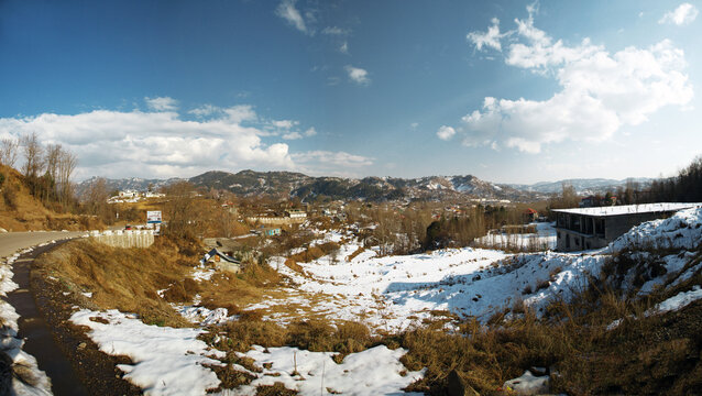 View of the snow-covered mountainous landscape with dry grass, distant peaks, and a building under a clear blue sky in Rawalakot, Kashmir, Pakistan.