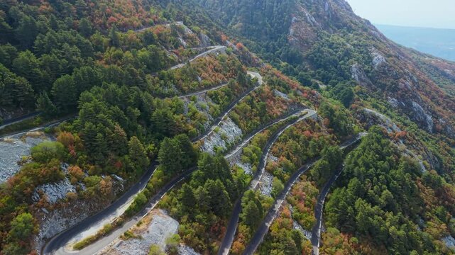Lovcen Serpentine Montenegro winds through forested mountain slopes in vivid autumn color. Aerial view follows tight road bends across rocky terrain, creating dramatic scenic route with calm grandeur.
