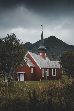 View of the historic red wooden Flakstad Church with its distinctive onion-shaped spire set against dramatic mountains under a moody cloudy sky in Ramberg, Nordland, Norway.