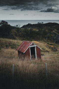View of the historic red wooden shed in Nesland surrounded by a grassy field and rocky coastal landscape in Flakstad Municipality, Nordland, Norway.
