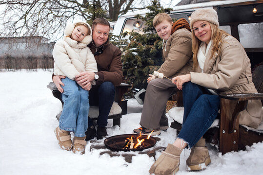 Family of four enjoying winter outdoors by a campfire, seated on wooden chairs with snow-covered ground and a vintage vehicle in the background