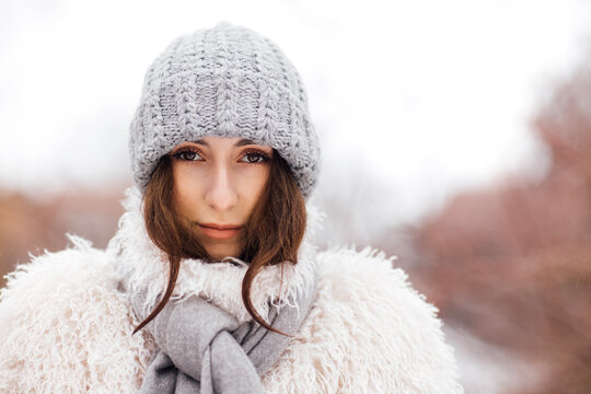 Young woman with long dark hair wearing a knitted gray beanie and a fluffy white coat poses outdoors in a snowy landscape with blurred trees in the background
