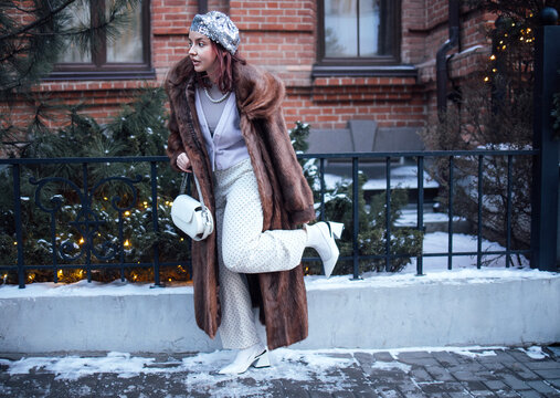 Fashionable young woman in fur coat and stylish hat poses on snowy street near brick building, showcasing winter outfit and accessories in urban setting