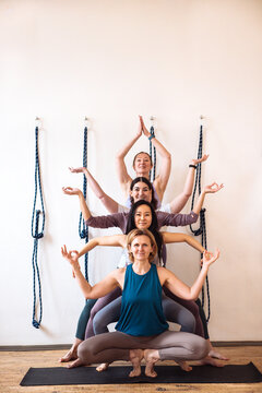 Group of four women practicing yoga in a stacked formation on mats, with yoga straps hanging on the wall in a bright studio during a wellness session
