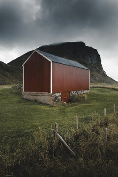 View of a red barn with a stone foundation set in a grassy field near Haukland Beach with a dramatic mountain in the background in Leknes, Nordland, Norway.