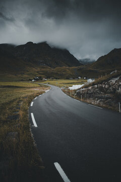 View of a winding asphalt road leading through a dramatic mountainous landscape under a moody, overcast sky near Haukland Beach in Leknes, Nordland, Norway.