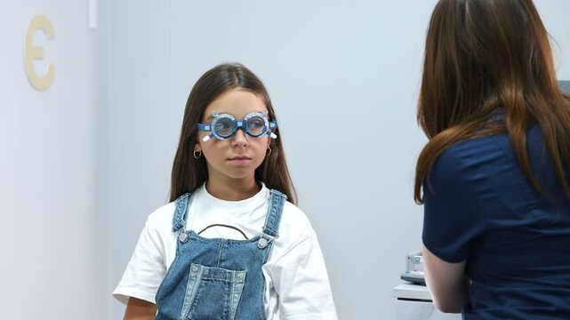 Female optometrist fits vision testing glasses on a young girl in a modern eye examination room, with medical equipment and tools in the background