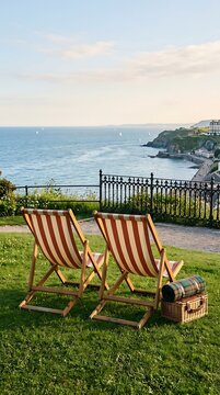 Two wooden lounge chairs on green grassy cliff overlooking distant coastal hotel scenery.