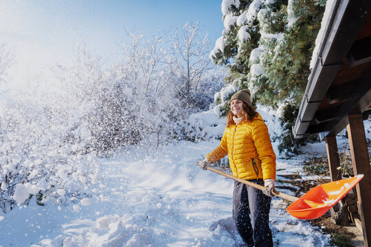 Happy caucasian woman smiling and shoveling snow near a country house terrace on a sunny winter day.