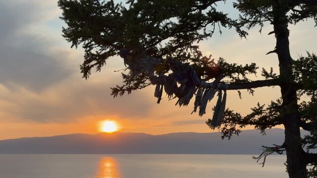 Video of Baikal Lake at sunset. View from shore of Olkhon Island to Small Sea against backdrop of setting sun. Colorful ribbons left by tourists and Buryat Hadaks flutter on branch of old larch tree.