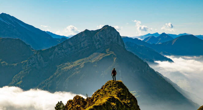 View of a person standing on a rocky peak in the Swiss Alps, overlooking a vast mountain range with clouds in the valleys under a clear blue sky in Airolo, Ticino, Switzerland.