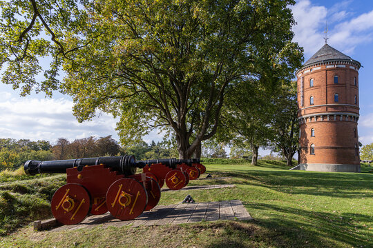 View of historic cannons on red carriages positioned on a grassy field near a brick water tower at Nyborg Castle, surrounded by large trees, Nyborg, Fyn, Denmark.