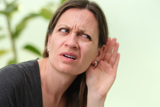 Close view of adult woman leaning forward with hand cupped beside ear, showing puzzled skeptical reaction in bright home interior with soft green plant backdrop and shallow focus