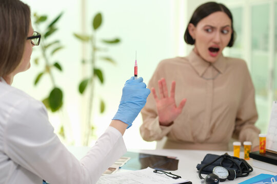 Gloved healthcare worker extends syringe toward startled woman in bright modern clinic exam room, conveying vaccination or treatment, patient anxiety, and careful medical attention