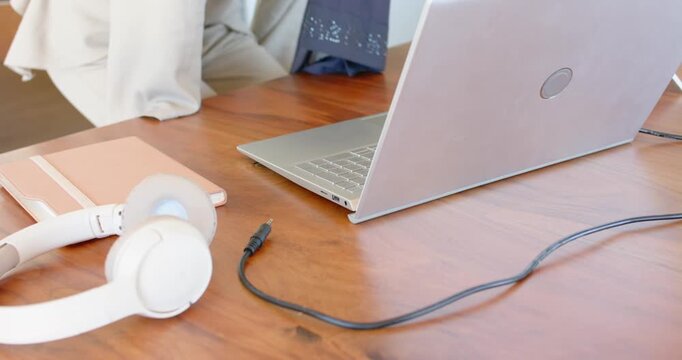 Woman approaching chair, sitting at work desk, wearing scarf, plugging cable into laptop to work