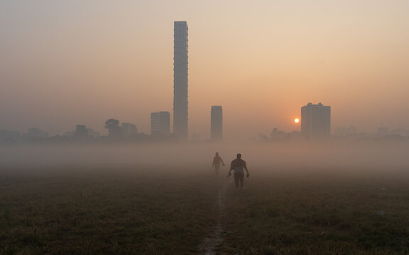 View of two people walking across a grassy field in the early morning fog with the city skyline and sunrise in the background in Kolkata, West Bengal, India.