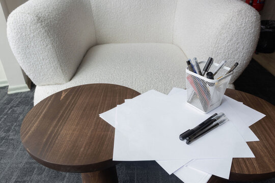 White armchair beside a wooden table with paper and pens. It represents a modern workspace and creative office environment.