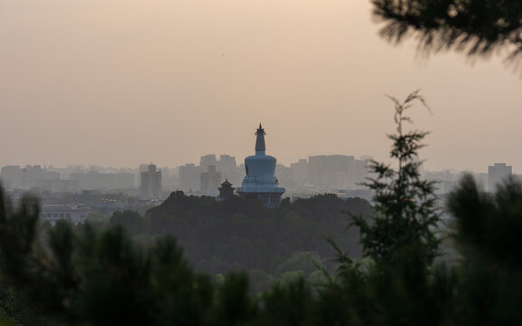 View of the White Dagoba in Beihai Park with the Beijing city skyline in the background framed by pine tree branches at sunset in Beijing, Beijing, China.