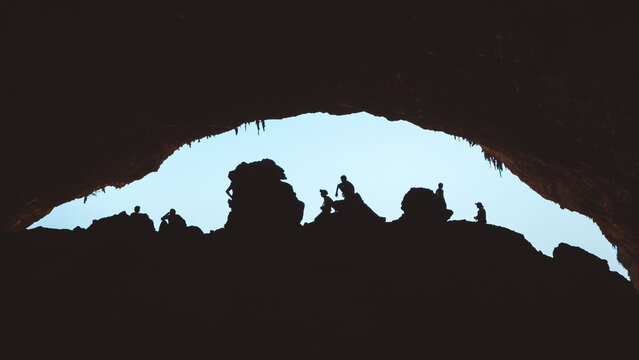 View of silhouetted people standing inside the large entrance of Hoq Cave, with rocky formations and bright sky visible in Socotra, Socotra, Yemen.