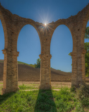 View of ancient stone arches with a sunburst shining through the structure, set against a rural landscape with plowed fields and a clear blue sky in Ponzano di Fermo, Marche, Italy.