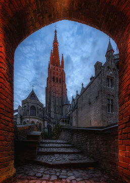 View of the Church of Our Lady framed by a historic brick archway with cobblestone steps leading towards the gothic tower in Bruges, Flanders, Belgium.