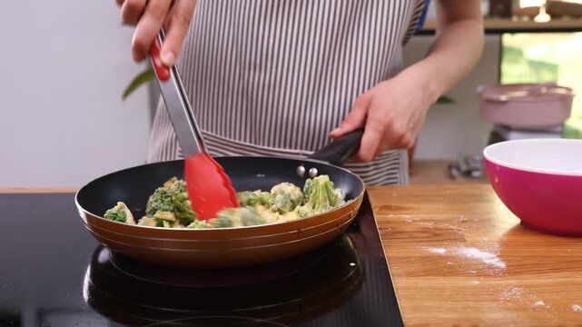 Housewife uses kitchen tongs to turn broccoli florets in hot frying pan. Woman prepares savory omelet enriched with nutritious vegetables time lapse