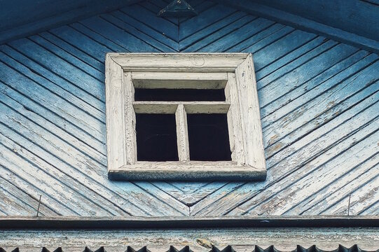 A small old white wooden window without glass in a wall of blue wooden planks in the attic of a country house