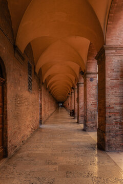 View of a long, historic brick arcade with vaulted ceilings and stone flooring, creating a symmetrical perspective leading into the distance in Urbino, Marche, Italy.