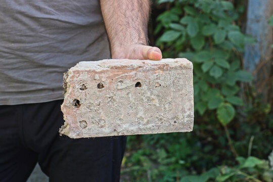 a man hand holds a old big brown gray one piece of brick on a summer street