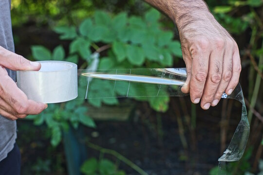 A mans hands hold a long strip of clear tape from a dispenser against a green background on a summer street