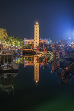 View of the lighthouse in the harbor of Fano at night, with fishing boats moored in the calm water reflecting the structure and the dark sky, Fano, Marche, Italy.