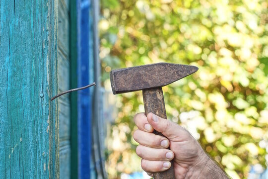 A one brown hammer in a workers hand drives a crooked nail into a green wooden board on a wall on a summer street