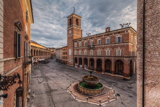 View of the historic Piazza del Comune with its central fountain, clock tower, and surrounding brick architecture in Fabriano, Marche, Italy.