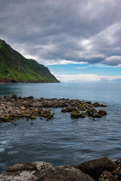 Povoa&ccedil;&atilde;o coastline showcasing volcanic rocks in the ocean foreground, leading to calm blue waters and lush green cliffs under a cloudy sky in S&atilde;o Miguel, Azores