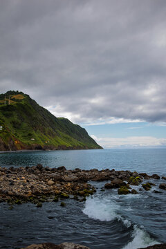 Atlantic Ocean waves crashing against the rugged Povoa&ccedil;&atilde;o coastline, revealing volcanic rocks and vibrant green hills under a cloudy sky in S&atilde;o Miguel, Azores