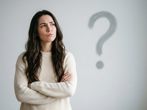 Confused young woman in beige knitted sweater stands with arms crossed looking towards hand painted grey question mark on wall as she ponders a difficult decision.