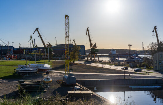 View of the industrial port area with large yellow cranes, docked sailboats on trailers, and a cargo ship in the background under a clear sky, Klaipeda, Klaipeda County, Lithuania.