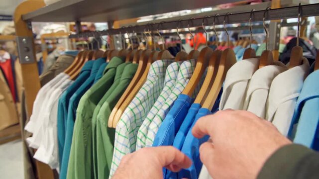 Close-up of a customer's hands browsing shirts and T-shirts on wooden hangers in a fashion store, comparing colors, patterns and fabrics while carefully choosing an item that suits personal style.