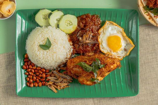 Traditional Malaysian nasi lemak served with fragrant coconut rice, spiced fried chicken, sunny side up egg, sambal, crispy anchovies, roasted peanuts and cucumber slices on a green plate. Classic Mal